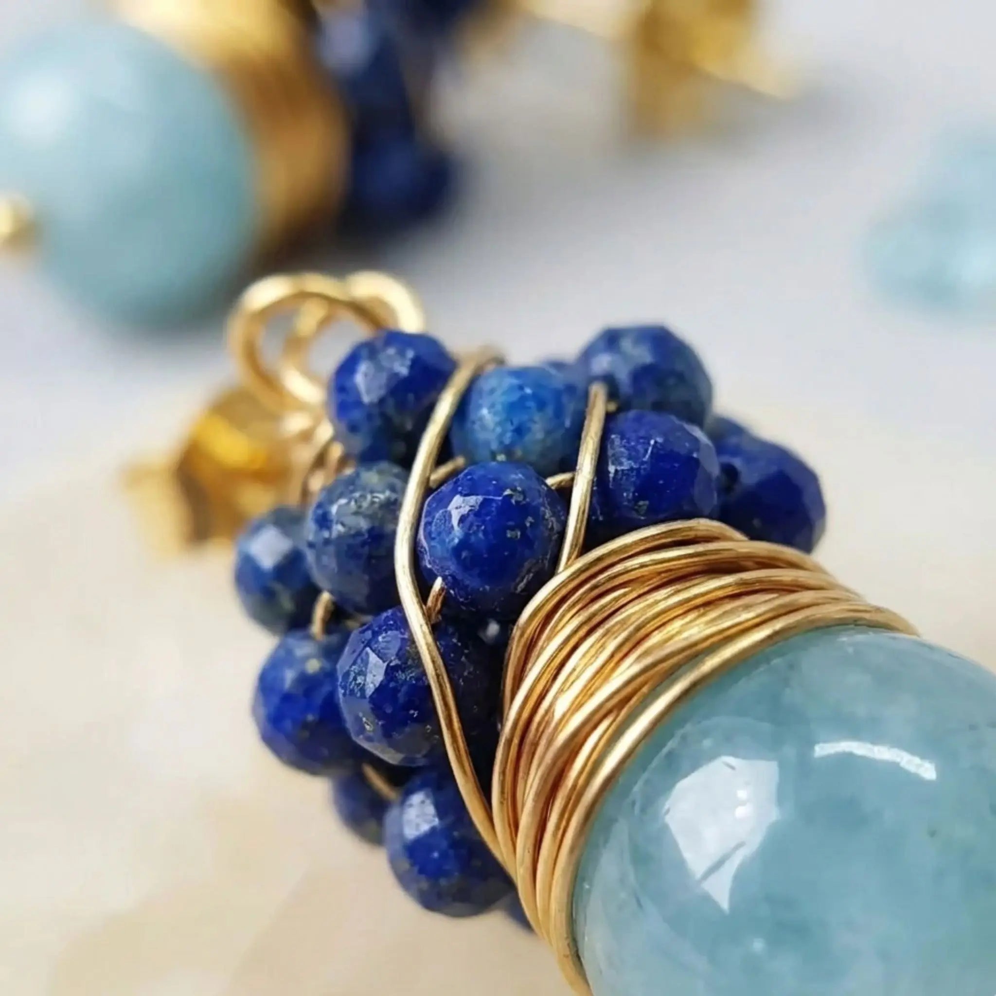 Très gros plan (macro) de haute qualité sur une boucle d'oreille Sanaca. L'image révèle la minutie du travail artisanal : le tissage complexe de fils d'or enserre parfaitement une grappe de perles de lapis-lazuli bleu profond, juste au-dessus de la transparence cristalline d'une perle d'aigue-marine. La mise au point est nette sur la texture des pierres et le fil d'or, montrant l'authenticité et la qualité des matériaux.