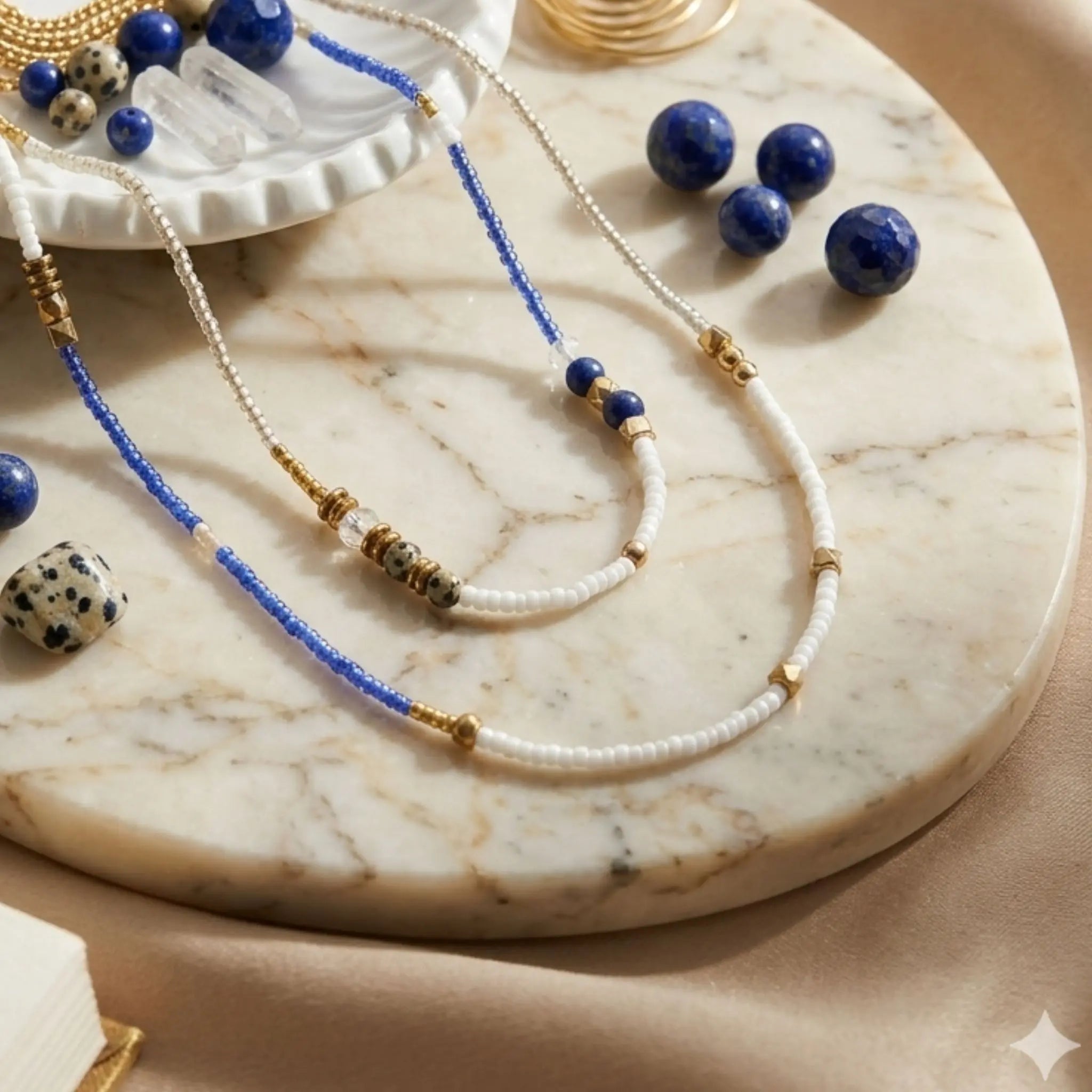 Vue rapprochée d'un collier sautoir artisanal en perles de Lapis-Lazuli bleues et Hématite dorée, disposé sur un plateau en marbre blanc avec des branches d'olivier séchées.
