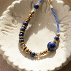 Gros plan détaillé d'une section du collier Marie dans une coupelle en céramique artisanale, montrant l'assemblage précis des perles de 6mm de Lapis-Lazuli, de l'hématite dorée facettée et des perles Miyuki 11.