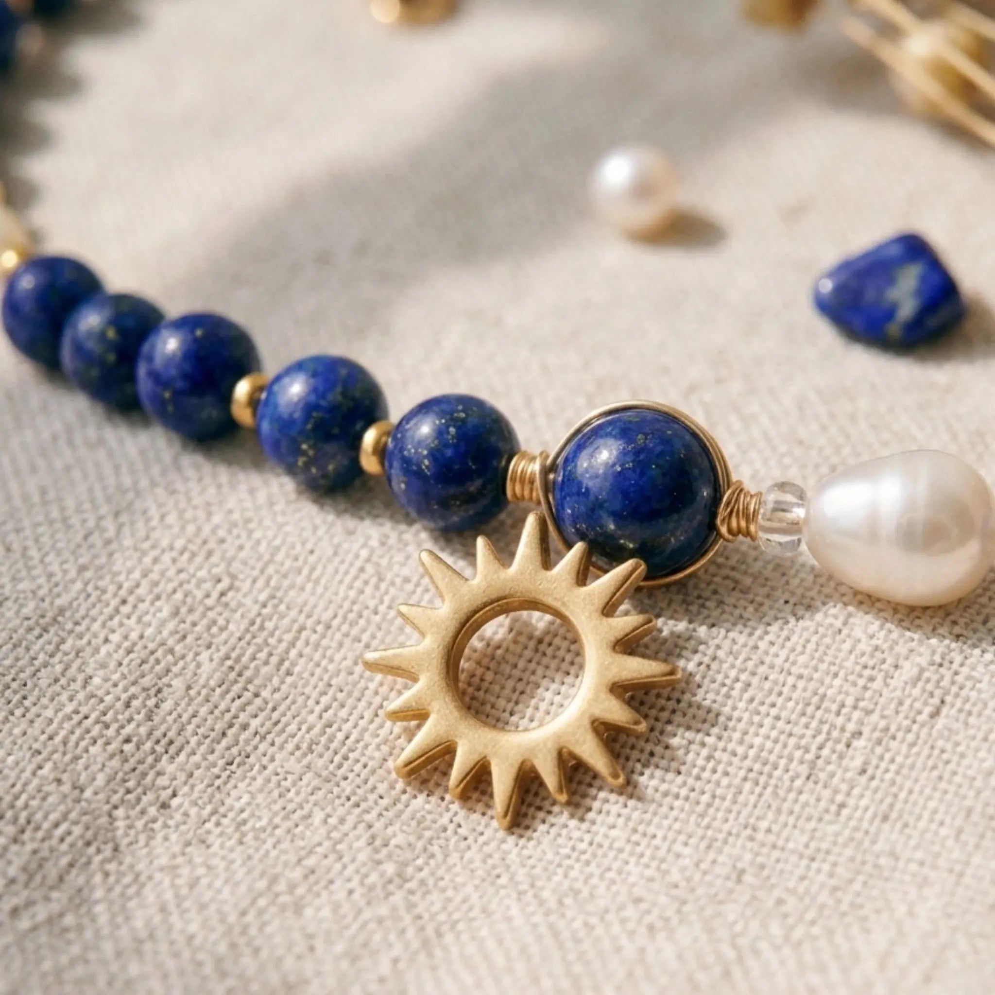 Vue macro du collier Solis montrant le pendentif soleil évidé en acier inoxydable doré à l'or fin. Le focus met en lumière les perles de lapis-lazuli bleu outremer de 6mm et une perle d'eau douce baroque nacrée, le tout assemblé à la main sur un tissu en lin beige naturel.