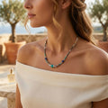 Plan rapproché d'une femme portant le collier Luz en ras-du-cou. Elle a une robe de lin blanc à épaules dénudées. On voit le collier Luz avec ses perles turquoises, bleues, transparentes et dorées reposant sur son cou. Le fond est un vignoble ensoleillé avec des oliviers et le logo doré SANACA sur la robe