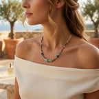 Plan rapproché d'une femme portant le collier Luz en ras-du-cou. Elle a une robe de lin blanc à épaules dénudées. On voit le collier Luz avec ses perles turquoises, bleues, transparentes et dorées reposant sur son cou. Le fond est un vignoble ensoleillé avec des oliviers et le logo doré SANACA sur la robe