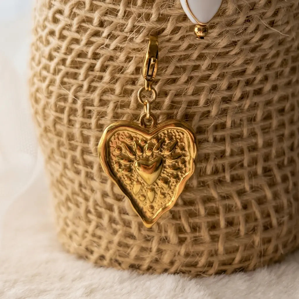 Un pendentif en acier inoxydable dore a l'or fin en forme de cœur texturé est suspendu à un anneau de saut en or sur un tissu de jute texturé. Le pendentif a un motif de soleil levant stylisé.