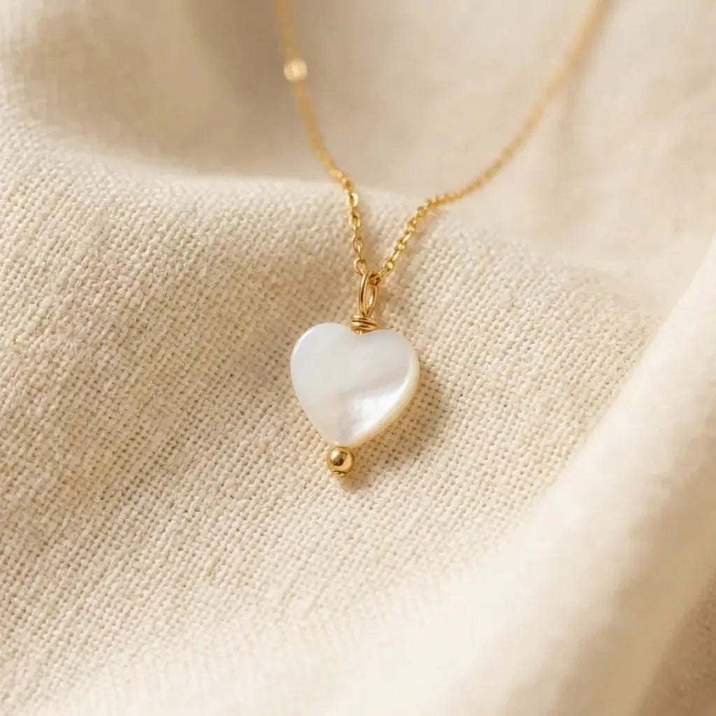 Un pendentif en forme de cœur en nacre blanche, suspendu à une fine chaîne en or, repose sur un tissu de lin beige texturé