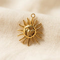 Gros plan d'un pendentif en acier inoxydable dore a l'or fin en forme de soleil au visage humain. Il repose sur un tissu de lin à armure lâche, doux et texturé. La breloque a un anneau de saut en haut.