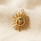 Gros plan d'un pendentif en acier inoxydable dore a l'or fin en forme de soleil au visage humain. Il repose sur un tissu de lin à armure lâche, doux et texturé. La breloque a un anneau de saut en haut.