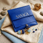 Une pochette en simili cuir bleu riche, texturée, avec le logo "SANACA" doré embossé sur le rabat. Posées délicatement devant, la paire de boucles d'oreilles en lapis-lazuli et aigue-marine. L'ensemble repose sur un assemblage de tissus en lin beige et bleu clair, entouré de galets de rivière et d'une gousse de graine séchée, créant une ambiance artisanale et naturelle pour la présentation du bijou.