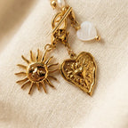 Une collection de breloques et de composants de bijoux en or suspendus ensemble sur un tissu de lin texturé. Les breloques comprennent un soleil avec un visage humain, un cœur texturé, un cœur en nacre et d'autres perles.